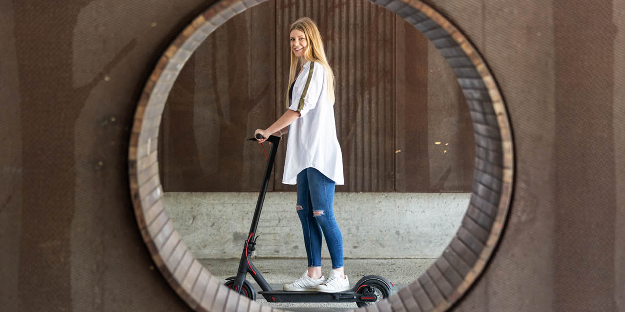 A woman stands on the scooter and smiles.